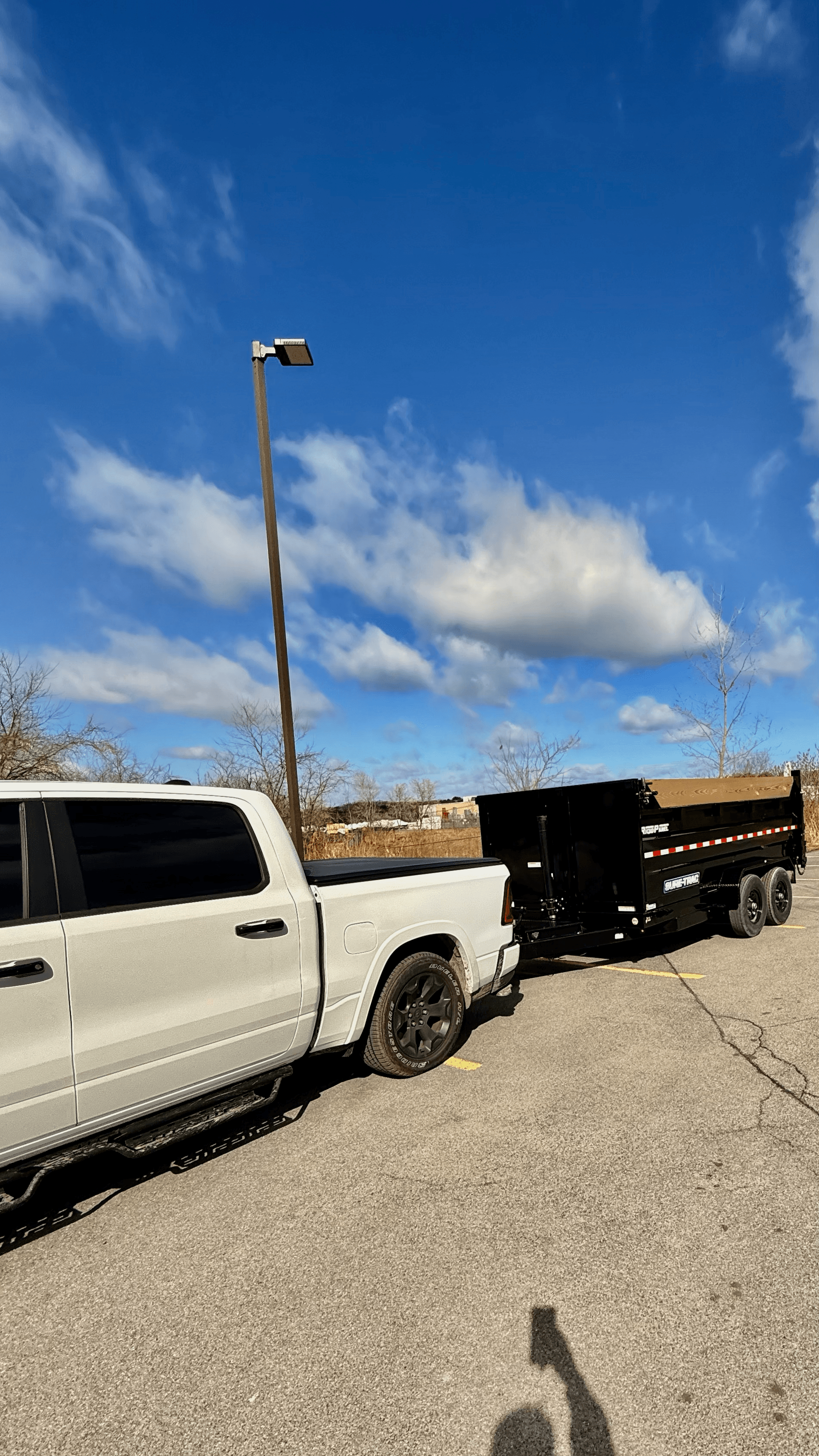 White pickup truck towing a black dump trailer on asphalt under a bright blue sky.